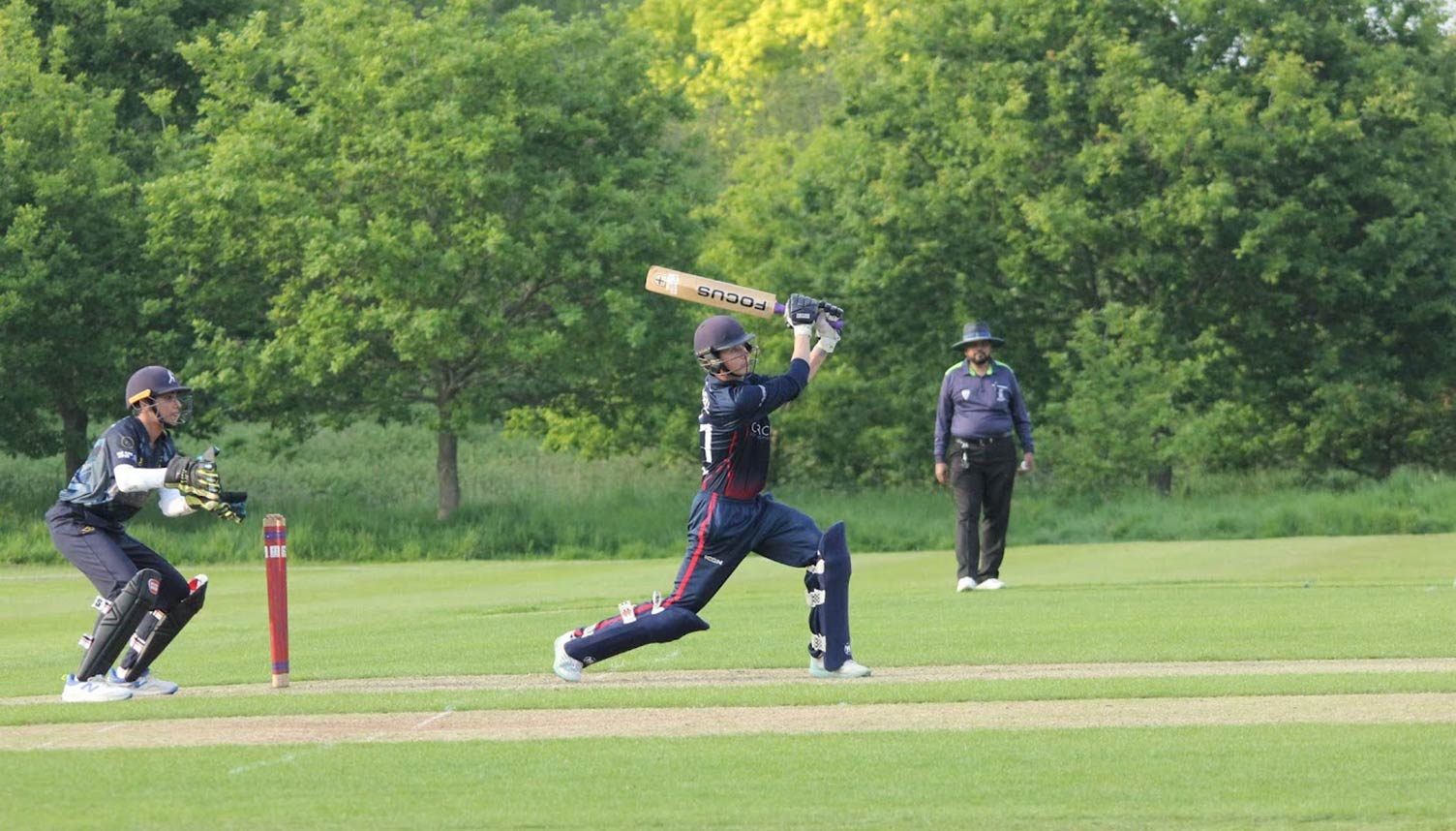 A batter playing an attacking drive on a cricket pitch. A wicket keeper is standing up to the stumps while an umpire looks on. In the background are trees lining the boundary.